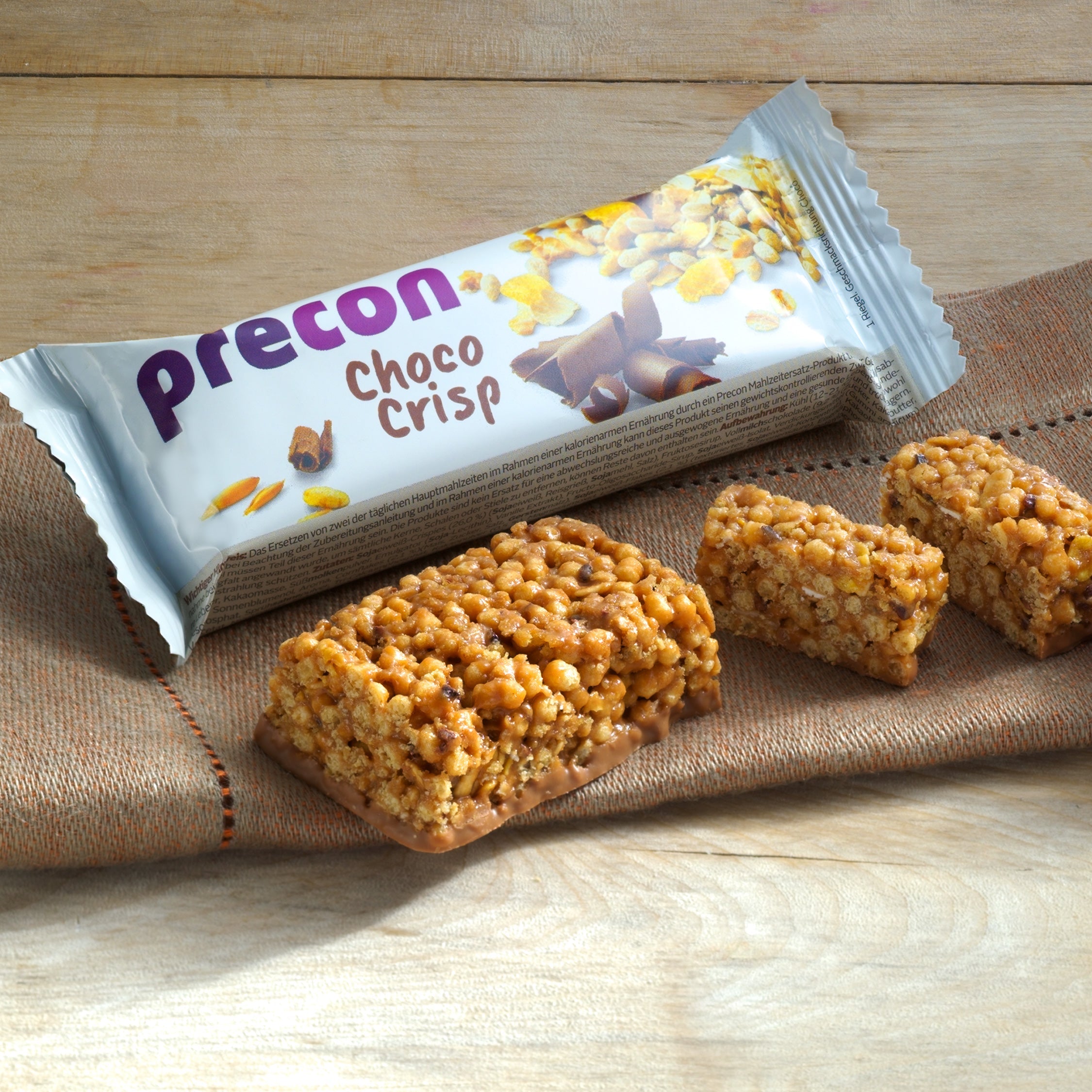 Ein verpackter Precon-Riegel aus dem 3-Monats-Mega-Vorrats-Paket CH wird auf einer Serviette mit drei unverpackten knusprigen Schokoriegeln auf einer Holzunterlage gezeigt - ideal für ein effektives Gewichtsmanagement.