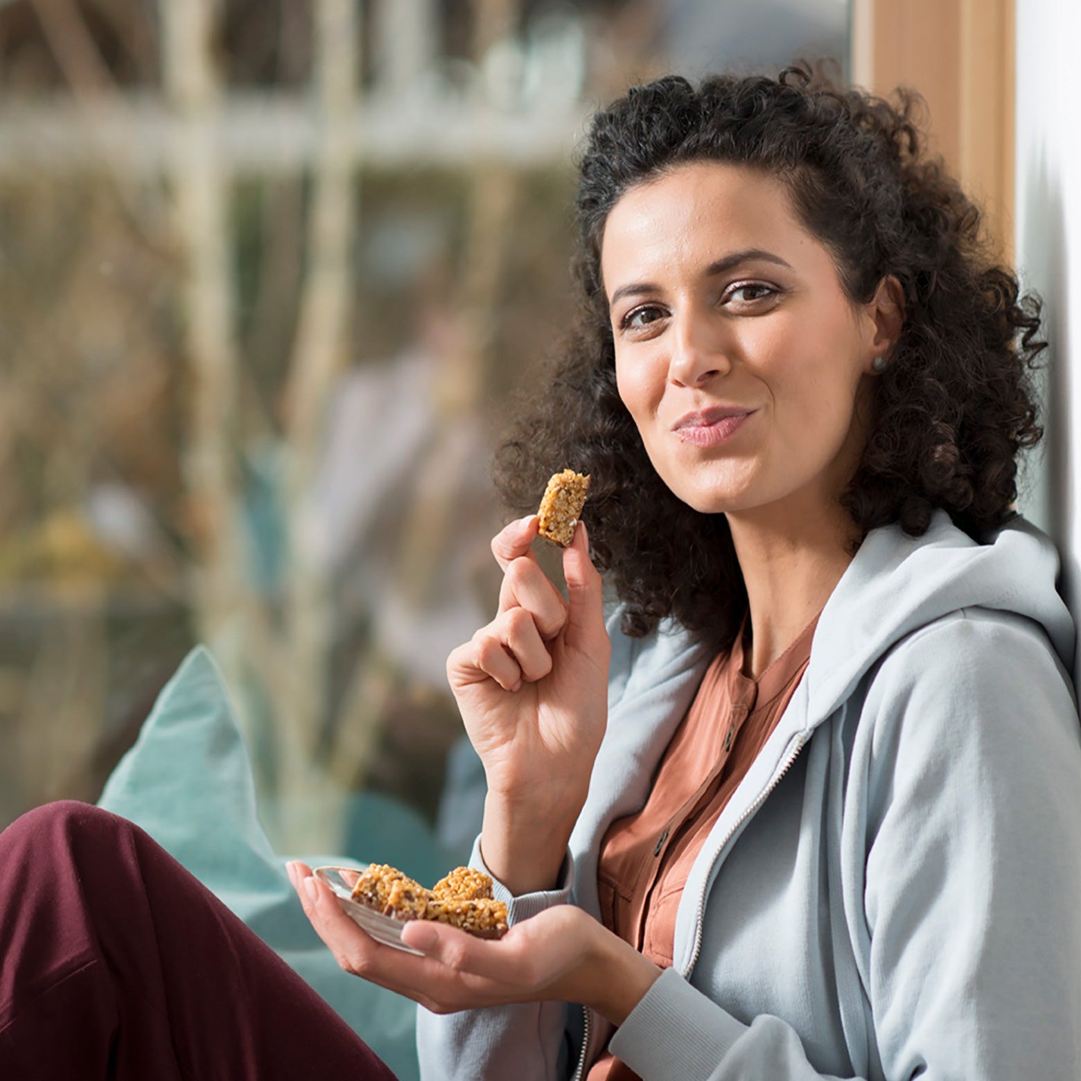Eine Frau mit lockigem Haar sitzt am Fenster, lächelt und hält einen Teller Precon ChocoCrisp Riegel in einer Hand, während sie mit der anderen isst. Sie ist lässig mit einem Kapuzenpulli bekleidet und wirkt während ihrer Diät zur Gewichtsabnahme entspannt und glücklich.
