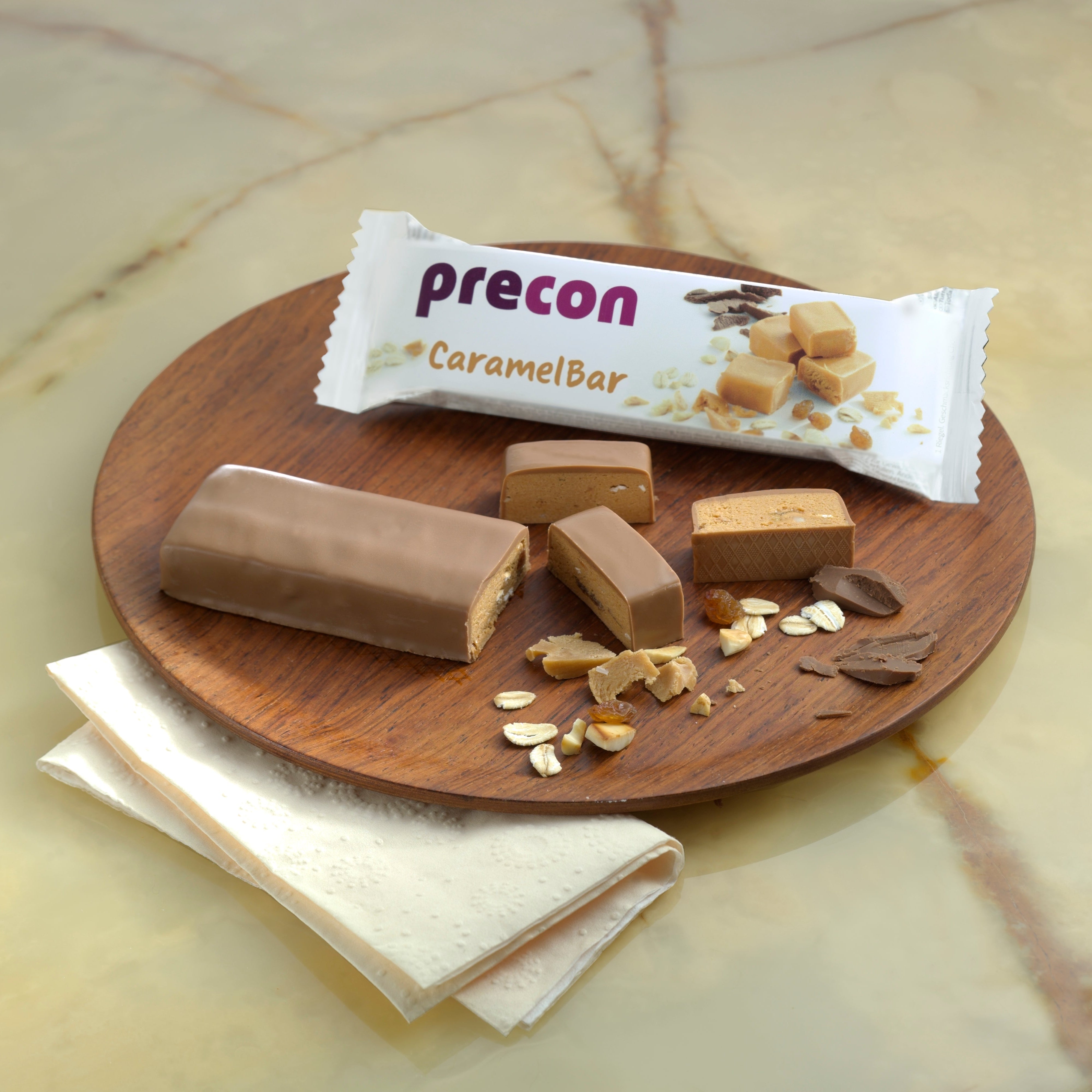 Auf einem Holzteller liegt ein Precon 3-Monats-Mega-Vorrats-Paket CH Karamellriegel, geschnittene Stücke, Haferflocken, Nüsse und Schokoladensplitter neben der weißen Verpackung und Servietten auf Marmor - eine köstliche Hilfe beim Gewichtsmanagement.