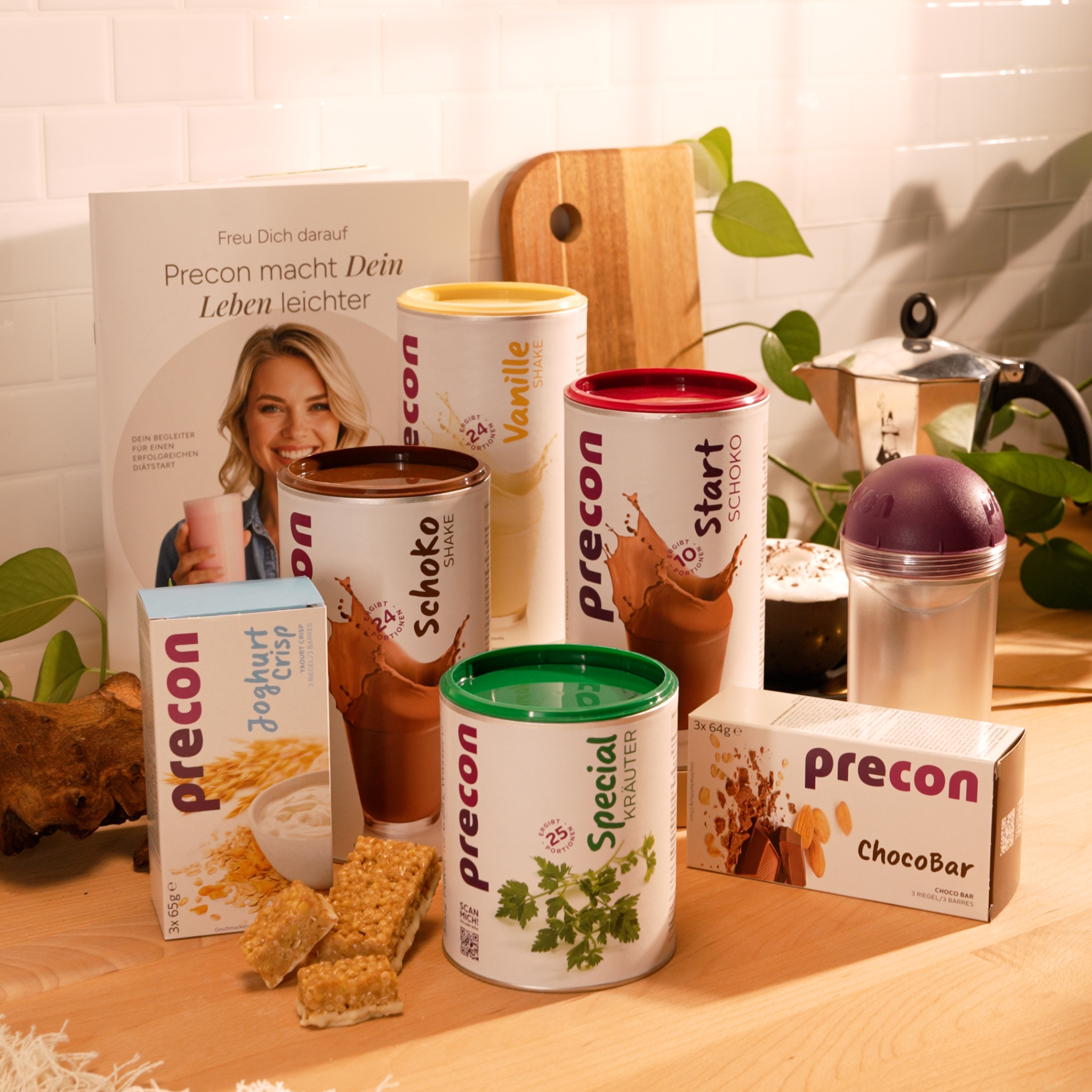Das Precon 4-Wochen-Neujahrspaket mit Getränkepulver, ChocoBars und einem Shaker steht auf einer Küchentheke mit einem Rezeptheft, einem Schneidebrett und einer Kaffeekanne im Hintergrund.