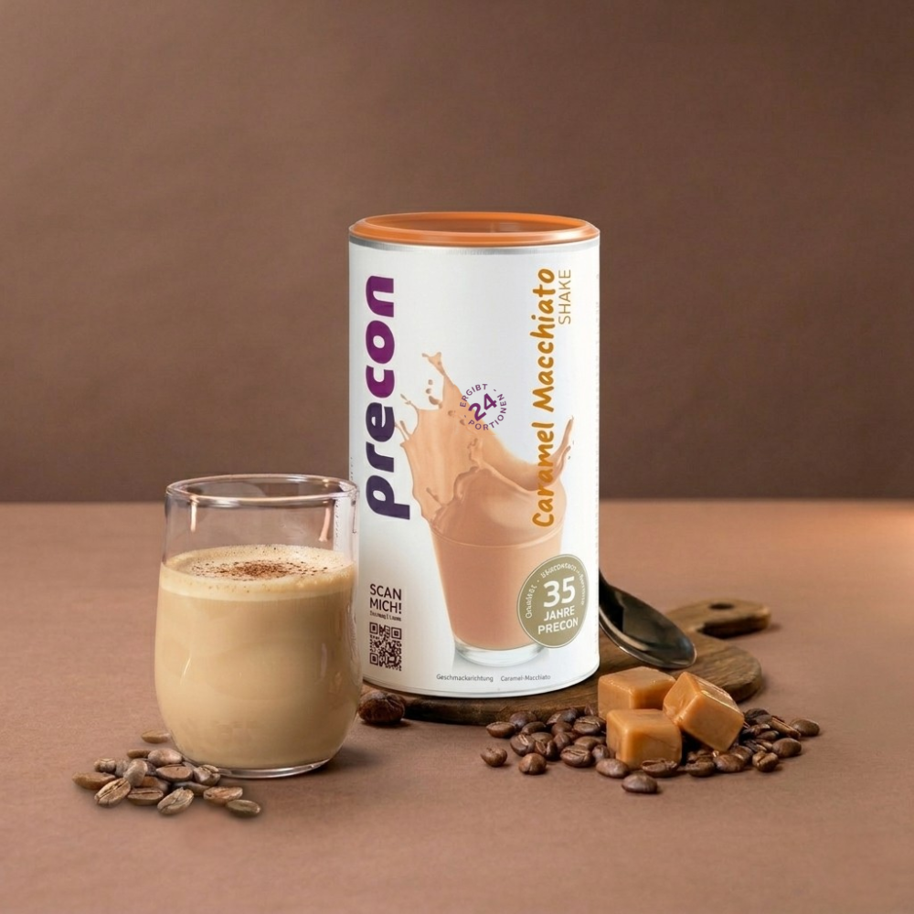 Ein Glas Caramel Macchiato Shake steht neben einem Kanister mit der Aufschrift Anona GmbH Caramel Macchiato Shake, umgeben von Kaffeebohnen und Karamellwürfeln auf einer braunen Fläche mit braunem Hintergrund.