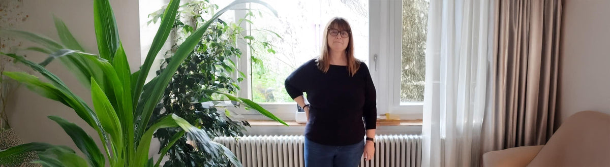 Corinne (59) nach ihrer Gewichtsabnahme mit Precon in ihrem Wohnzimmer neben einer großen Pflanze