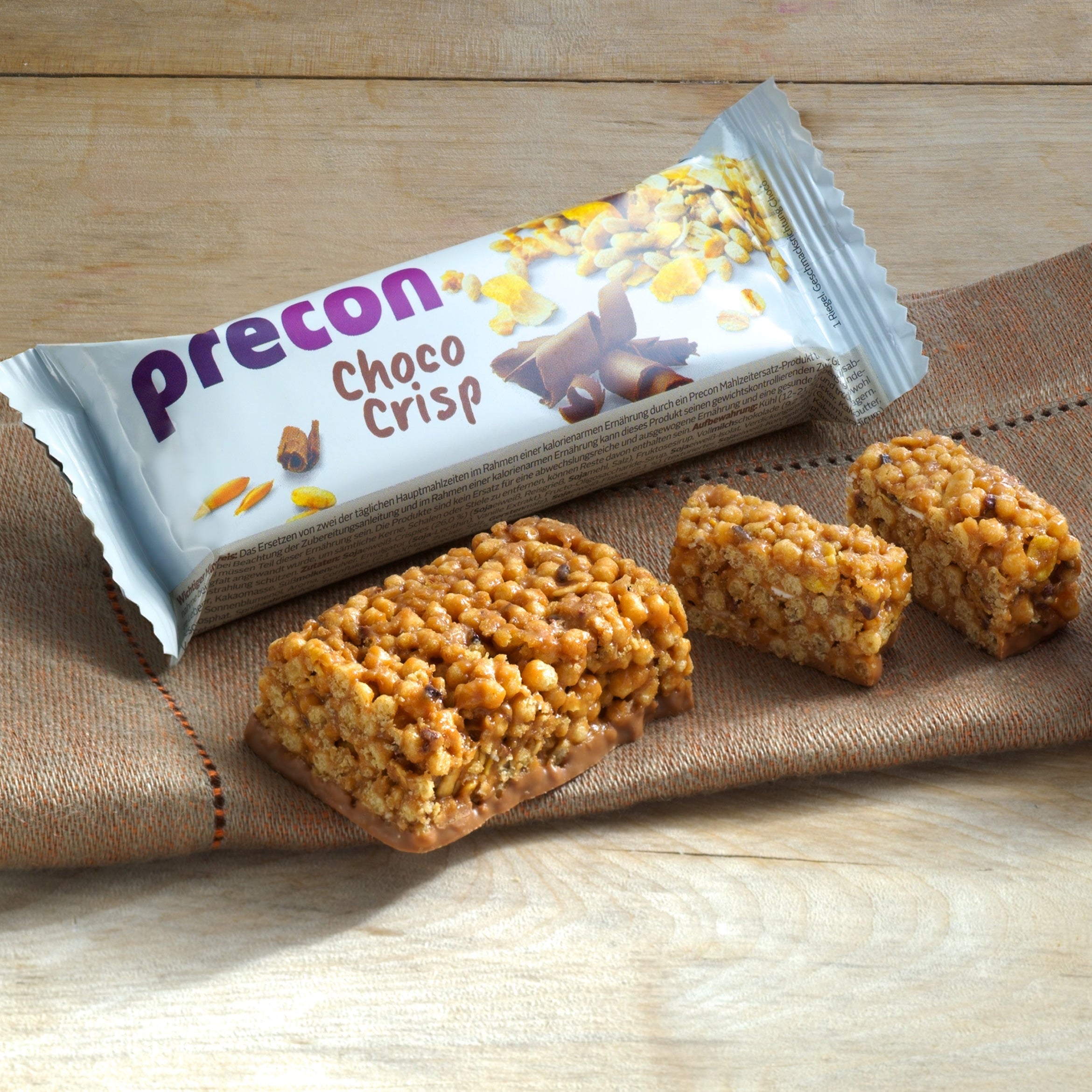 Ein verpackter Precon Black Friday 4-Wochen-Paket-Riegel liegt auf einer Holzfläche neben drei unverpackten knusprigen Snack-Riegeln, die auf einem braunen Tuch liegen.
