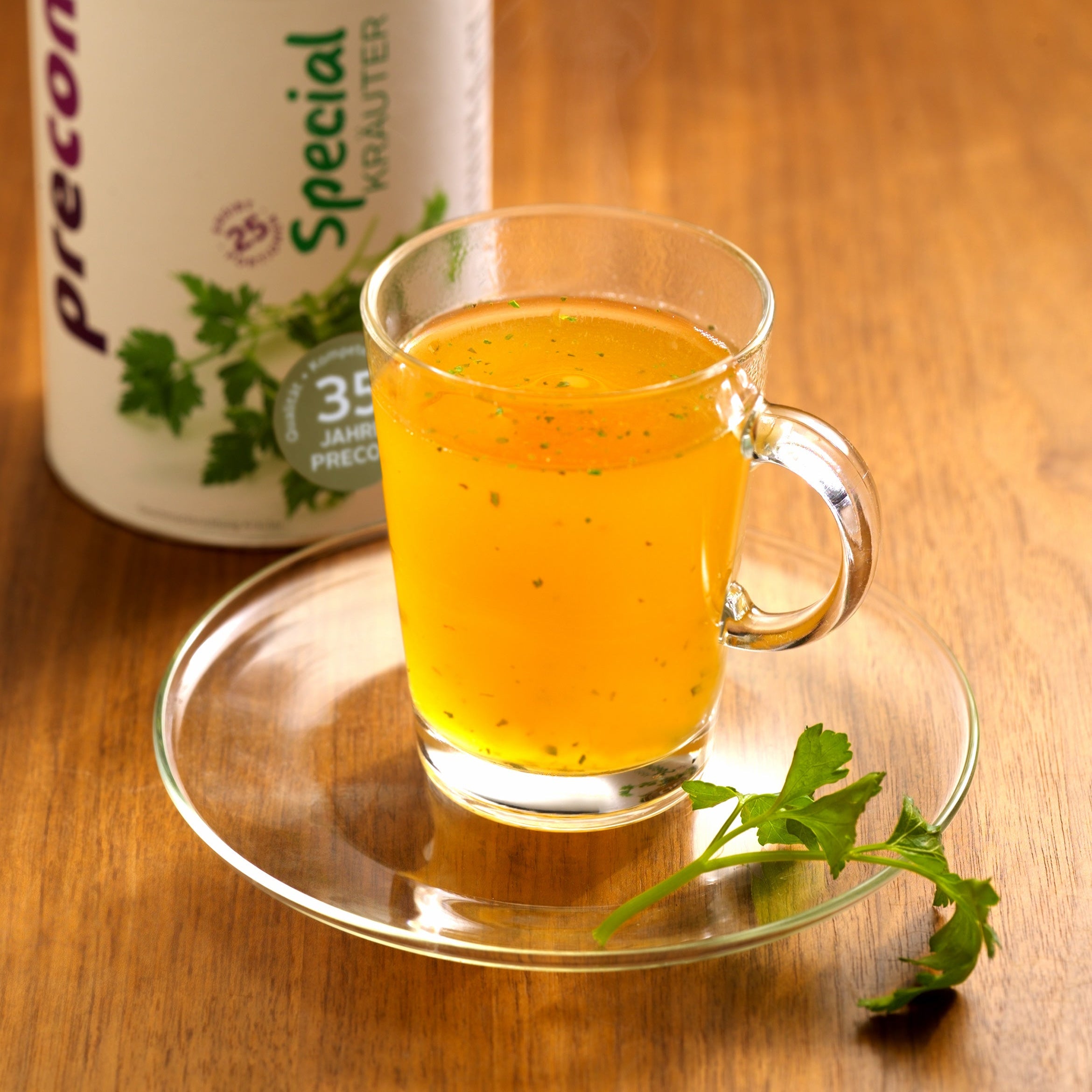 Ein Klarglasbecher mit gelbem Special Kräuter steht auf einer Untertasse, daneben liegt frische Petersilie. Im Hintergrund ist teilweise ein großer Precon Special Kräuter Behälter für Diät und Fettverbrennung zu sehen.