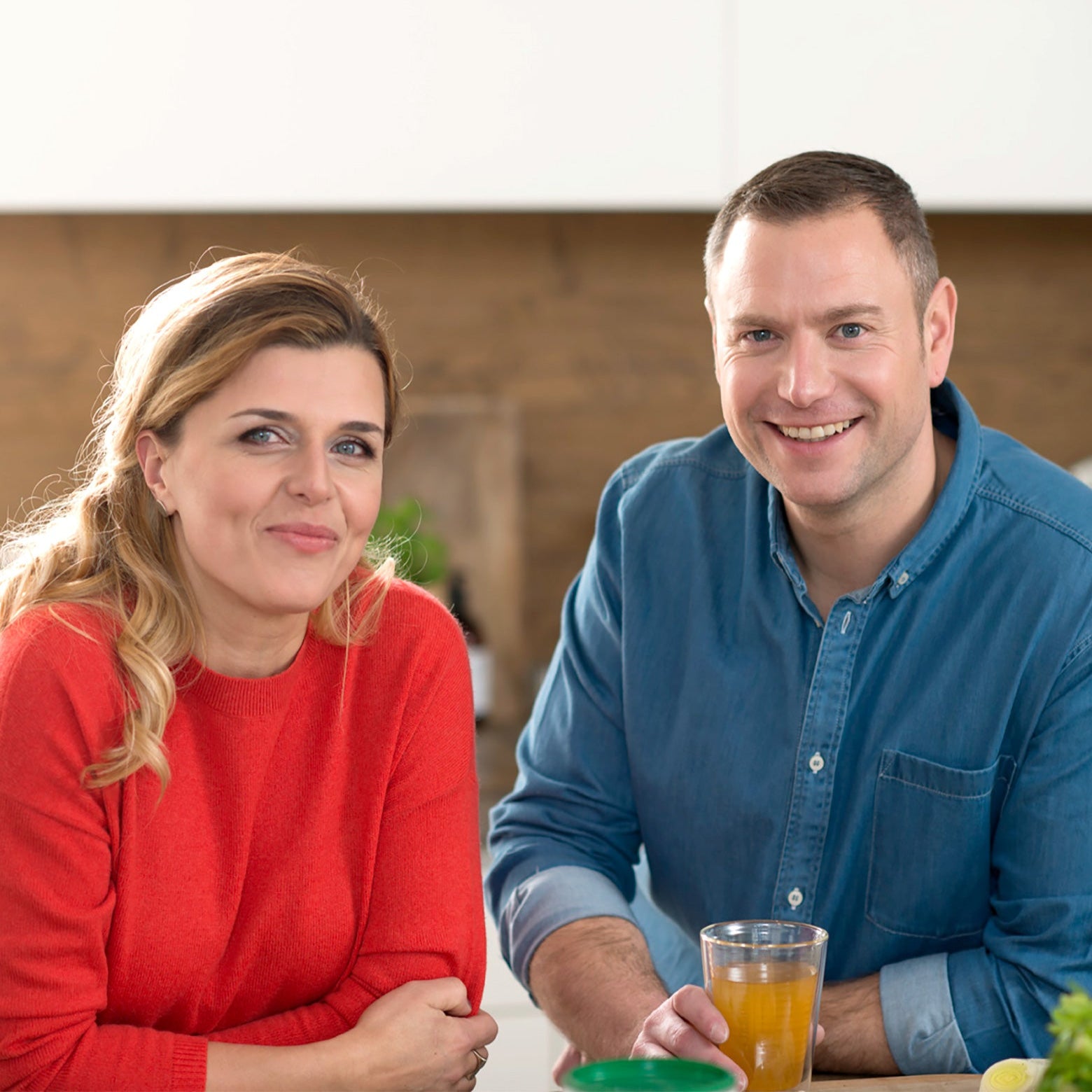 Eine Frau in einem roten Pullover und ein Mann in einem blauen Hemd lächeln an ihrem Küchentisch. Er hält Orangensaft in der Hand, der ideal für ihre Ernährung ist, während Precon Special Kräuter in der Nähe steht, das perfekte Kräuterheißgetränk für ihren modernen Lebensstil.