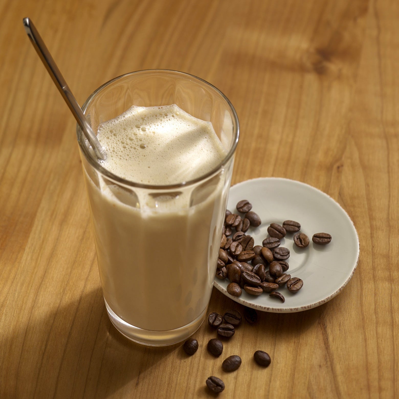 Ein Glas Eiskaffee mit Löffel steht neben einer Untertasse mit gerösteten Kaffeebohnen auf einem Holztisch - ein idealer Genuss während Ihres Precon 3-Monats-Mega-Vorrats-Paket CH für Diät- oder Gewichtsmanagement.