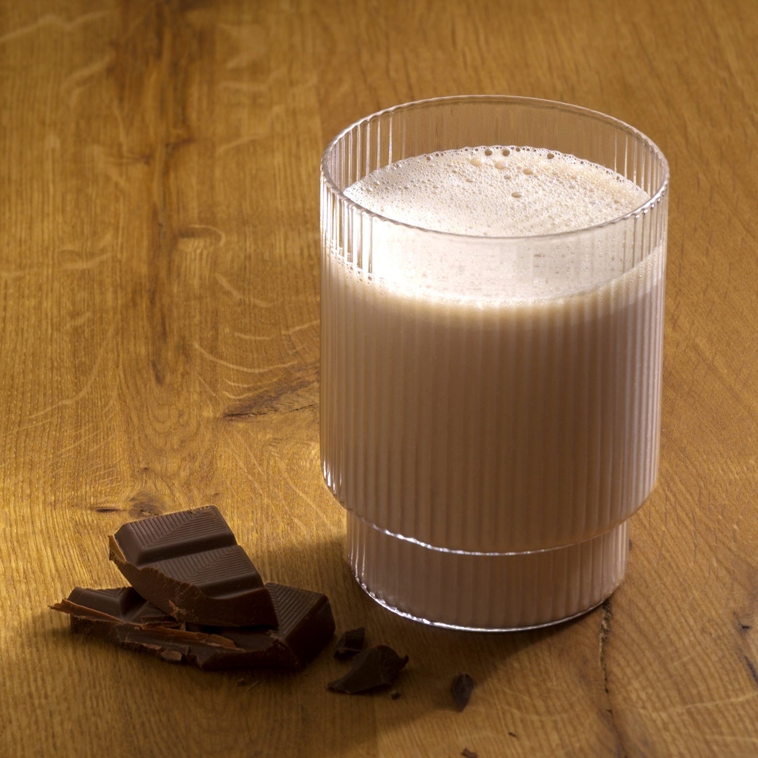 Ein Glas schaumige Schokoladenmilch aus dem Precon 3-Monats-Mega-Vorrats-Paket CH steht auf einer Holzplatte mit Schokoladenstückchen - perfekt als köstlicher Genuss während der Precon Diät für ein intelligentes Gewichtsmanagement.