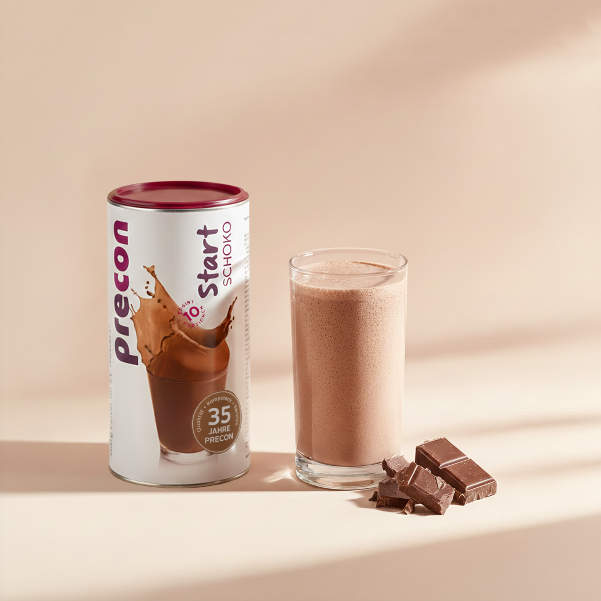 Ein Precon Start Shake Schoko-Behälter steht neben einem Glas Schoko-Shake, daneben liegen Schokoladenstücke, alles vor einem beigen Hintergrund.