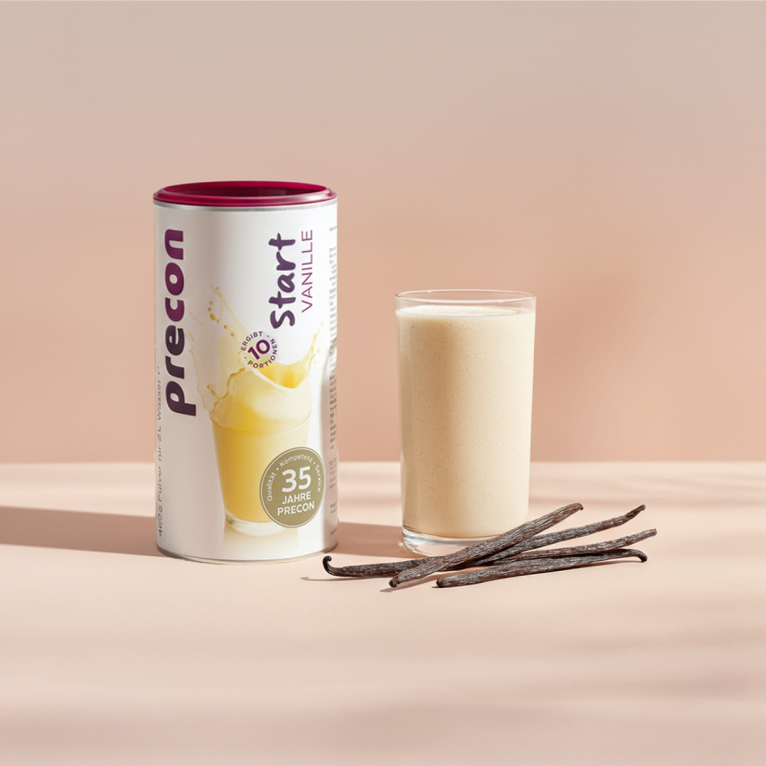 Ein Kanister Precon Start Shake Vanille wird neben einem gefüllten Glas und drei Vanilleschoten auf einer hellen Fläche vor einem neutralen Hintergrund aufgestellt.