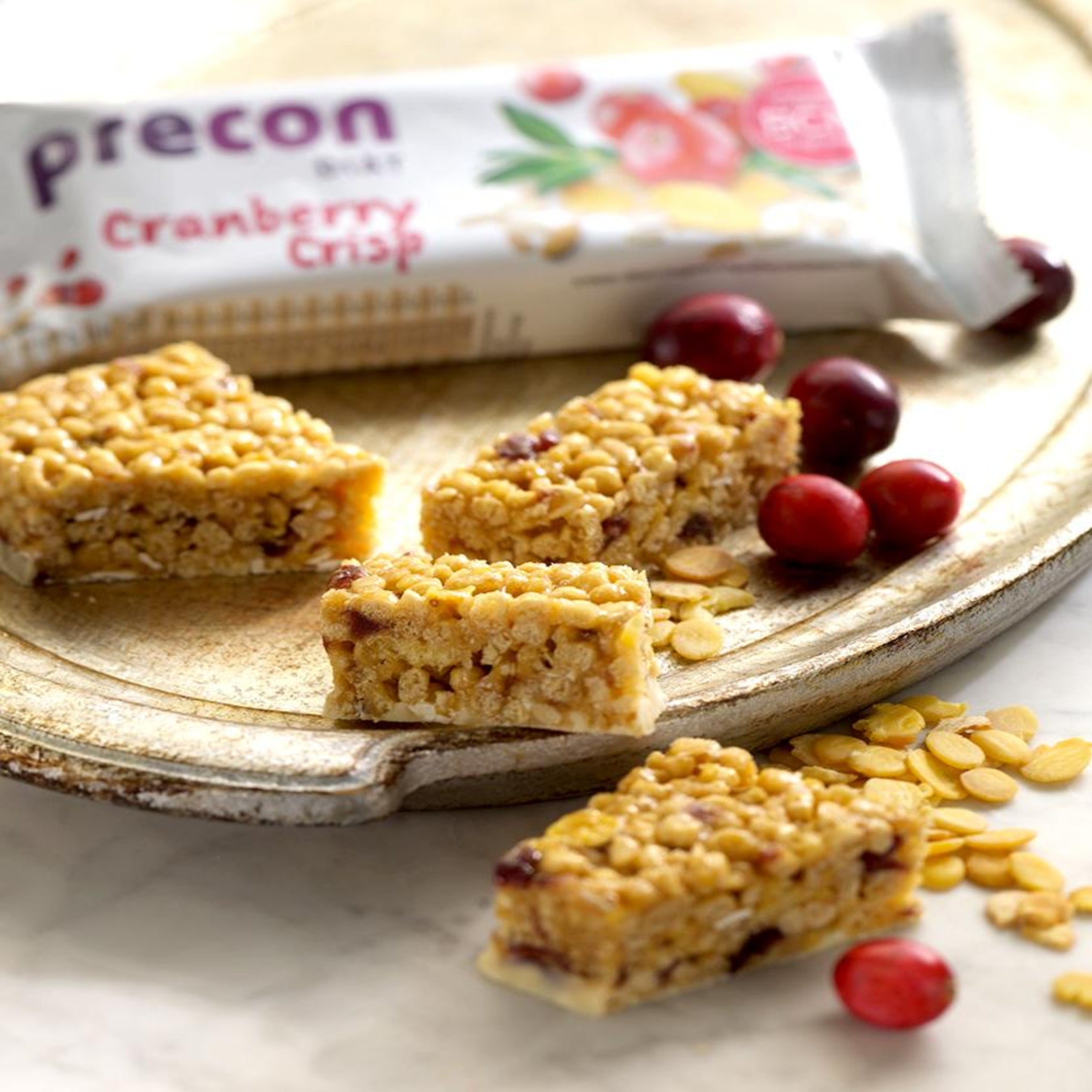 Rechteckige Müsliriegel mit Cranberries liegen auf einem Keramikteller, umgeben von Hafer und Cranberries. Im Hintergrund ist ein teilweise geöffnetes Precon 2-Monats-Vorrats-Paket Cranberry-Crisp-Verpackung für effektiven Gewichtsverlust.